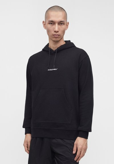 Άνδρας με ξυρισμένο κεφάλι που φοράει μαύρο φούτερ με τσέπη μπροστά και το λογότυπο "A-COLD-WALL*" στο κέντρο του στήθους, στέκεται απέναντι σε απλό φόντο.