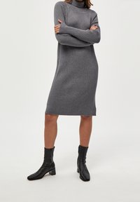 Femme portant une robe en maille côtelée gris à col roulé et des bottes noires à talons mi-mollet, debout les bras croisés devant un fond blanc.