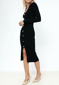 Robe noire à côtes avec un décolleté en V plongeant, manches longues, détails de boutons à l'avant et fente sur le côté, associée à des escarpins nus.