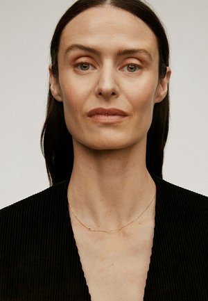 Collar de oro con un pequeño acento, usado sobre piel desnuda. La modelo tiene el cabello lacio y oscuro, y viste una blusa negra con escote en V profundo.