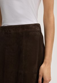 Bruine suède rok met een gladde textuur, voorzien van voornaaddetails en een verborgen ritssluiting aan de zijkant. Gecombineerd met een witte top.