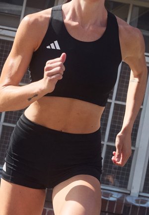 Lekkoatleta w czarnym sportowym staniku i spodenkach Adidas biegnie na zewnątrz przed budynkiem z oknami w stylu kratownicy.