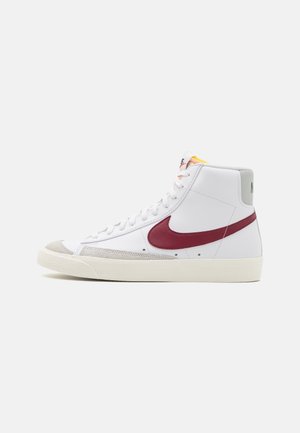 Biały wysokiego typu sneaker z bordowym logo Nike, beżowym zamszowym noskiem i kremową podeszwą, pokazany z profilu na jednolitym tle.