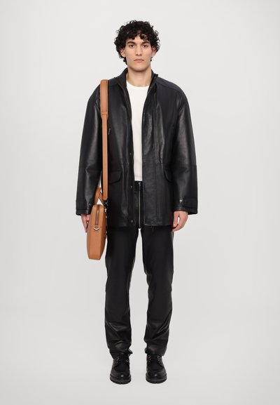 Chaqueta de cuero negra con dos bolsillos delanteros, llevada sobre una camisa blanca. Pantalones negros y botas negras, sosteniendo un bolso de hombro color marrón.