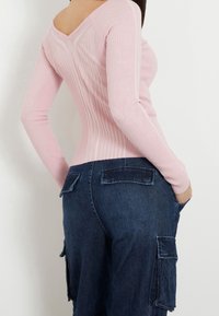Žena má na sobě růžový žebrovaný top s dlouhým rukávem a V-výstřihem na zádech, spolu s tmavě modrými džínovými cargo kalhotami s klopovými kapsami.
