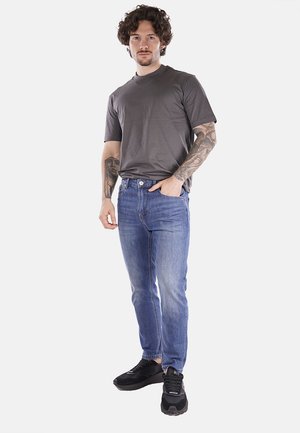 T-shirt grigio a maniche corte, jeans in denim blu, sneaker scuri. I jeans presentano una vestibilità classica e un leggero sbiadimento. Il modello sta in piedi con le mani nelle tasche.