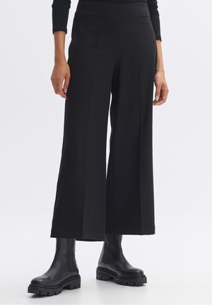 Femme portant un pantalon noir à jambes larges et coupé au-dessus de la cheville, associé à des bottines en cuir noir épaisses sur fond blanc.