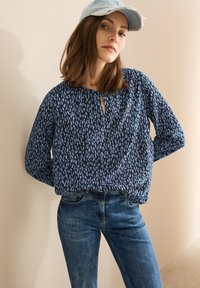 Blusa blu navy con un motivo testurizzato di linee verticali blu chiaro, con scollatura a chiave e vestibilità comoda. Abbinata a jeans blu.