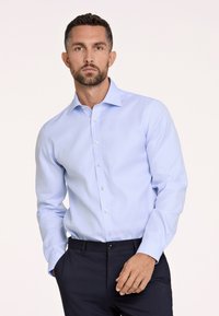 Camicia azzurra chiara con colletto a punta, maniche lunghe e chiusura con bottoni. Tessuto liscio con un leggero lucido. Indossata con pantaloni scuri.