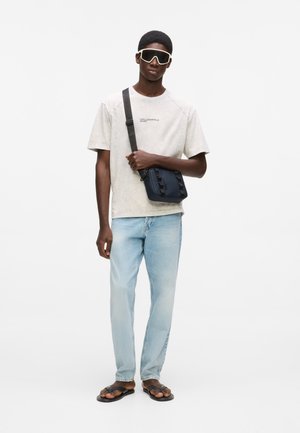 Jonge man draagt een oversized licht beige Karl Lagerfeld T-shirt, lichtblauwe spijkerbroek, zwarte sandalen, grote witte zonnebril en een donkerblauwe crossbody tas.