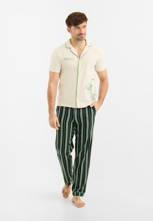 Mężczyzna w kremowej koszuli z krótkim rękawem na guziki z motywem panterki oraz w ciemnozielonych, pasiastych spodniach, chodzący boso na białym tle.