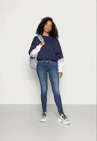 Sweatshirt azul-marinho oversized com detalhes em roxo e branco, combinado com jeans skinny azuis e ténis brancos. Leva uma bolsa azul-clara.