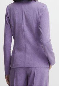 Lila kavaj i texturerat tyg, med skräddarsydd passform, långa ärmar och synliga sömmar, visad från baksidan.