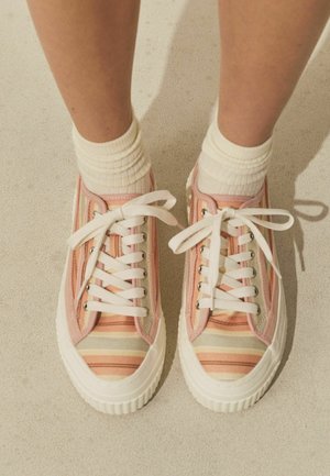 Nohy v bielych ponožkách a ružových plátených teniskách s pruhmi a bielymi šnúrkami, stojace na svetlej podlahe.