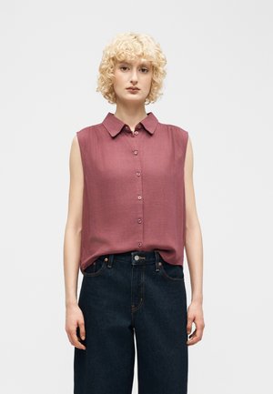 Žena s krátkymi kučeravými blond vlasmi, oblečená v bezrukávovej fialovo-ružovej košeli so zapínaním na gombíky, zastrčenej do vysokých tmavomodrých džínsov.