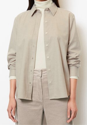 Kobieta ubrana w beżową koszulę zapinaną na guziki na biały golf i beżowe spodnie sztruksowe, stojąca na jednolitym białym tle.