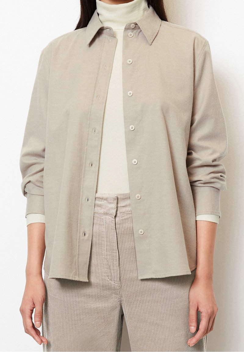 Femme portant une chemise beige boutonnée par-dessus un col roulé blanc et un pantalon beige en velours côtelé, debout devant un fond blanc uni.