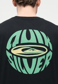 Άτομο που φορά μαύρο πουκάμισο με μεγάλο κυκλικό λογότυπο Quiksilver σε πράσινο και κίτρινο στην πλάτη, ορατό από πίσω και μέχρι τον λαιμό.