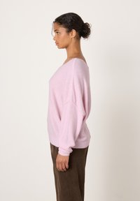 Moteris rožinės spalvos laisvu megztiniu ir rudomis korduro kelnėmis stovi profilio pozoje priešais paprastą šviesų foną.