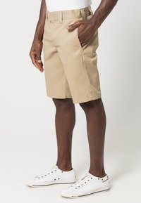 Beige bomullsshorts med rak skärning, sidofickor och en mjuk textur, parat med vita sneakers, mot en neutral bakgrund.