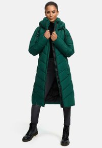 Cappotto lungo verde a puffo con cappuccio, caratterizzato da una cerniera frontale, cuciture a chevron e una finitura leggermente lucida, abbinato a stivali neri.