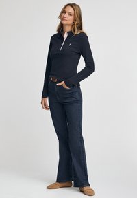 Marineblaues Langarm-Poloshirt mit rosa Kragen und Knopfleiste, kombiniert mit dunkelblauen, ausgestellten Jeans und hellbraunen Mokassin-Schuhen.