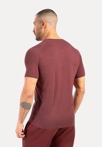 Man med kort hår och blommig tatuering på underarmen, iklädd marsala-färgad kortärmad skjorta och matchande byxor, vänd bort från kameran mot en neutral bakgrund.