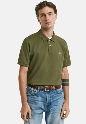 Olivgrünes Poloshirt mit klassischem Kragen, zwei-Knopf-Leiste und kleinem Logo. Kombiniert mit hellblauen Jeans und einem braunen geflochtenen Gürtel.
