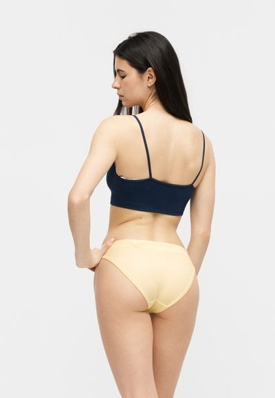 Γυναίκα με μακριά σκουρόχρωμα μαλλιά, οραστή από πίσω, που φοράει μπλε κομψό κορμάκι και ανοιχτό κίτρινο εσώρουχο, σε ανοιχτόφωτο φόντο.