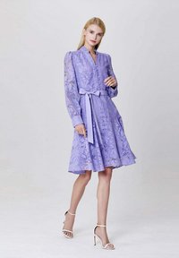 Robe violet clair avec de longues manches transparentes, motif floral, boutonnière à l'avant, ceinture à la taille et jupe évasée. Le mannequin porte des talons blancs à brides.