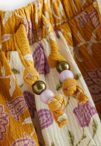 Ceinture à cordon avec des lacets en tissu jaune moutarde ornés d'un imprimé floral violet et de perles décoratives violettes et bronze sur un tissu texturé.