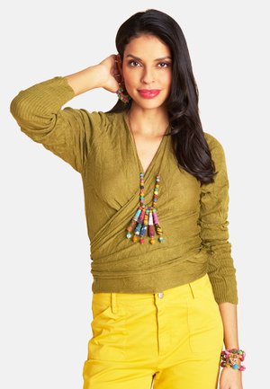 Frau mit langem dunklem Haar, die einen olivfarbenen Wickelpullover, leuchtend gelbe Hosen und eine bunte Perlenkette, Ohrringe und Armbänder trägt, posiert mit der Hand hinter dem Kopf.