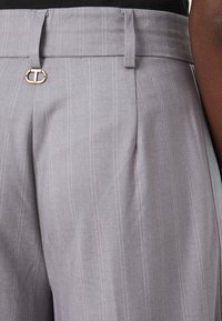 Pantalon gris clair en tissu lisse avec des rayures verticales. Présente un accent en quincaillerie dorée et une taille structurée avec des passants de ceinture.