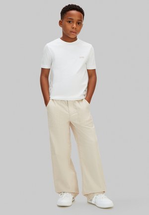 Niño de pie con las manos en los bolsillos, vistiendo una camiseta blanca de Calvin Klein, pantalones beige de pierna ancha y zapatillas blancas sobre un fondo gris claro.