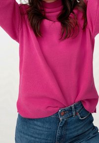 Pull tricoté fuchsia avec un col montant montant et une texture côtelée, associé à un jean bleu taille haute. Présente une coupe décontractée.