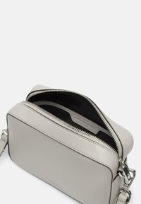 Bolso bandolera de cuero gris claro con cierre de cremallera, correa ajustable y forro interior negro. Presenta costuras sutiles y un detalle del logo.
