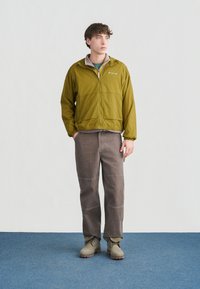 Νέος άνδρας που στέκεται με τα χέρια στις τσέπες, φορώντας πράσινο σακάκι Columbia, γκρι παντελόνι και μπεζ μποτάκια, σε μπλε χαλί φόντο.