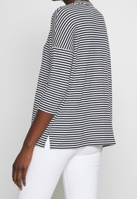 Maglia a righe bianco e nere a maniche lunghe con spacchi laterali, scollo rotondo e vestibilità rilassata. Il tessuto appare morbido e leggermente texturizzato.
