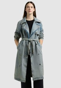 Donna che indossa un lungo trench in denim blu slavato con cintura sopra un abito nero, mani in tasca, in piedi davanti a uno sfondo grigio chiaro uniforme.