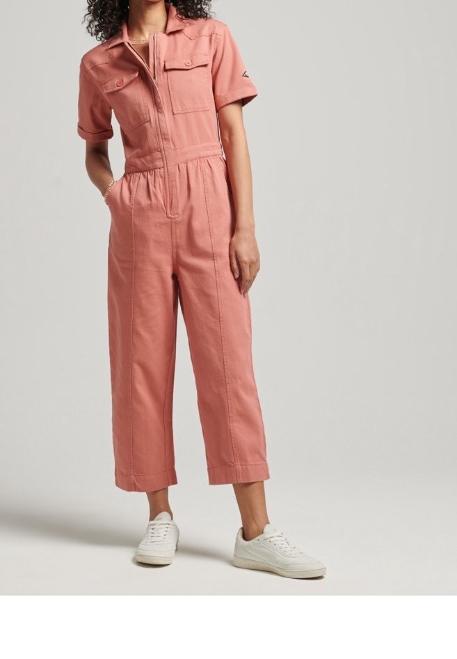 Różowy kombinezon z krótkim rękawem, dwoma kieszeniami na piersiach, elastyczną talią, krótkimi nogawkami i zamkiem błyskawicznym z przodu. Noszony z białymi trampkami.