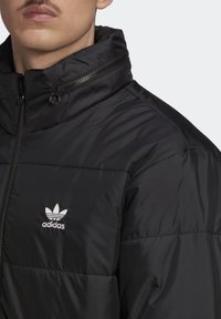 Czarna puchowa kurtka z wysokim kołnierzem, srebrnym logo adidas po lewej stronie, zamkiem błyskawicznym i gładką, błyszczącą fakturą.