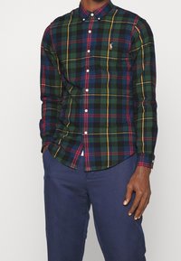 Zielona, niebieska i czerwona koszula z długim rękawem w kratkę z kołnierzykiem zapinanym na guziki i białymi guzikami. Posiada małe haftowane logo na klatce piersiowej.