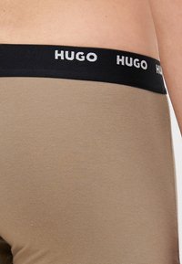 Beige Boxershorts aus weichem Stoff mit einem schwarzen elastischen Bund, der das "HUGO"-Logo in Weiß trägt. Glatte Textur, figurbetontes Design.