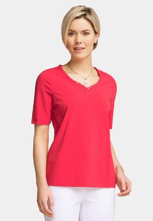 T-shirt en coton rouge avec un col en V et des manches courtes, présentant des clous en argent décoratifs le long de l'encolure. Tissu texturé, coupe décontractée.
