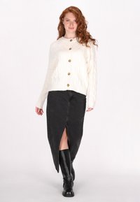 Crème kabelgebreide cardigan met gouden knopen, gecombineerd met een zwarte spijkerrok met een hoge split, gedragen met zwarte kniekousen.