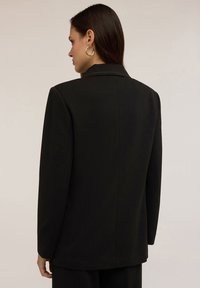 Blazer nero con una silhouette strutturata, revers a punta e uno spacco singolo sul retro, realizzato in un tessuto liscio e testurizzato.