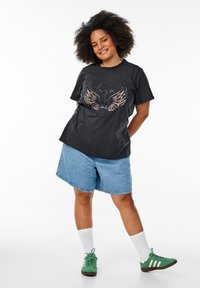 Camiseta de algodón negra con diseño gráfico de alas; combinada con pantalones cortos de denim azul claro. El modelo lleva zapatillas verdes y calcetines blancos.