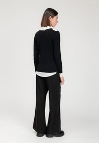Schwarzer Strickpullover mit weißem Kragen und goldenen Akzenten, kombiniert mit schwarzen weiten Hosen und schwarzen Plateauschuhen.