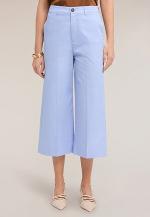 Kobieta nosząca pastelowo niebieskie, szerokie, skrócone spodnie z przodu zapinane na guzik i zamek, w połączeniu z beżowymi, ostro zakończonymi szpilkami i złotymi bransoletkami na lewej ręce.