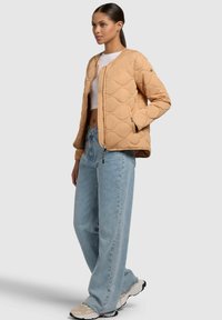 Giacca bomber beige trapuntata con zip frontale e polsi a costine, abbinata a jeans a gamba larga blu chiaro e sneakers neutre.